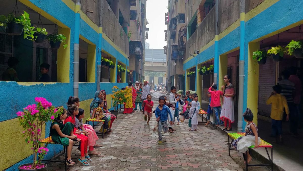 Children running in an alleyway between two buildings. Alleyway has benches, flower pots and a recent coat of paint. People of all ages are gathered in this space